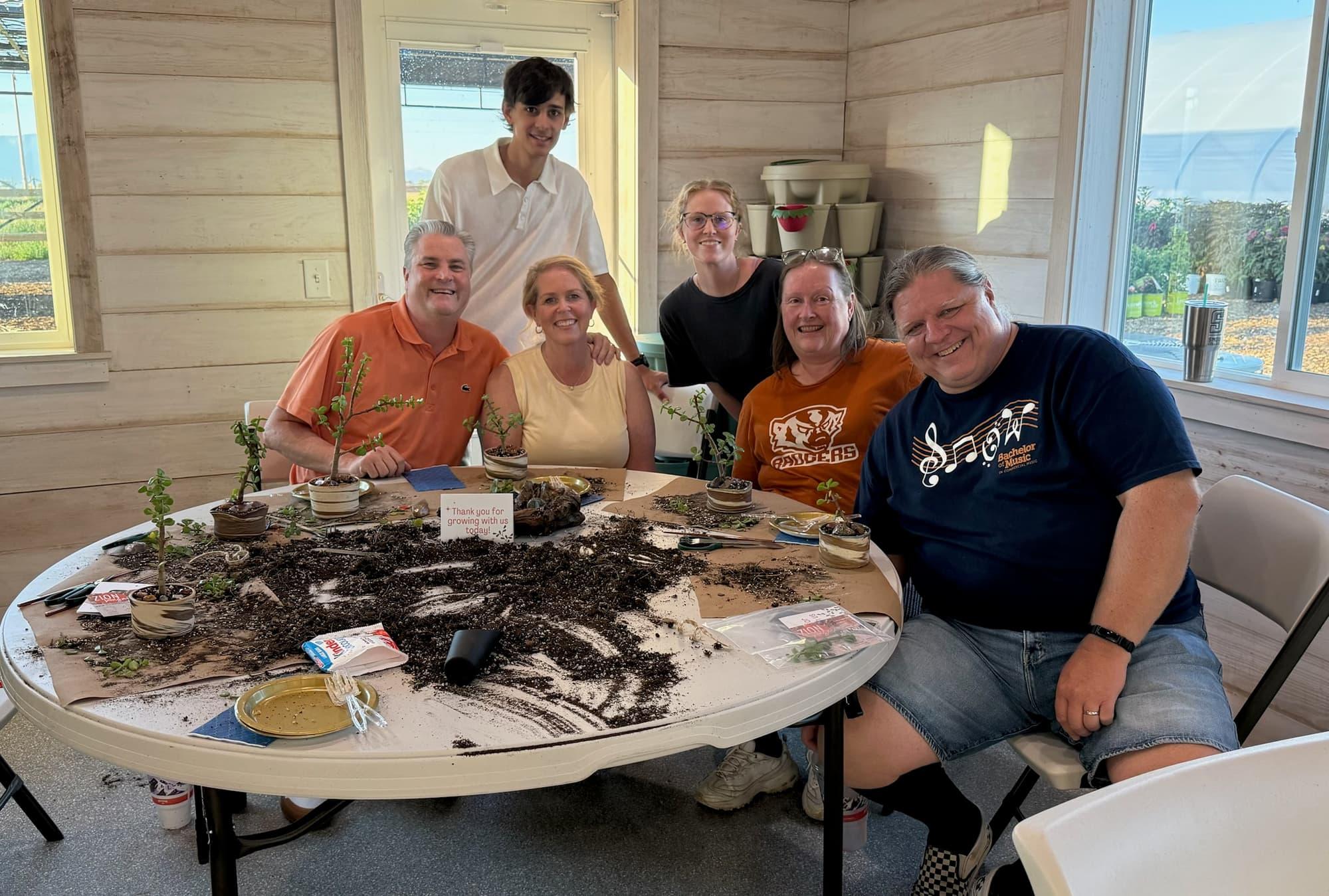 Six participants smile behind a round table covered in soil, small dwarf jade bonsai in front of them