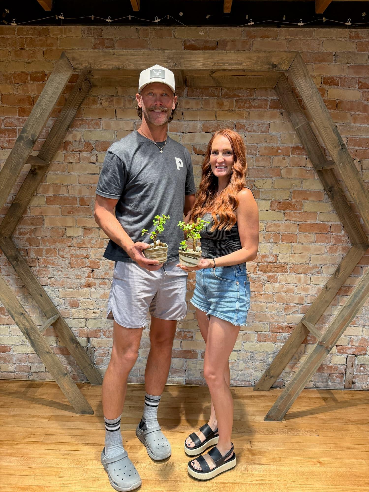 Two participants hold their finished dwarf jade bonsai in handmade pots in front of a brick wall